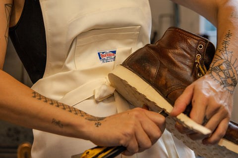 Close-up shot of the Rosa Rugosa Close Apron in Natural, with model Lo mending a shoe. 
