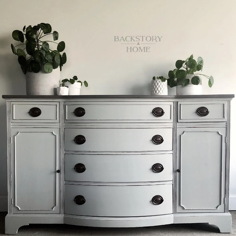 a vanity that has been painted in a French Grey with black/dark grey accents on the raised detailing and knobs. styled with indoor plants in white pots, against a neutral beige wall