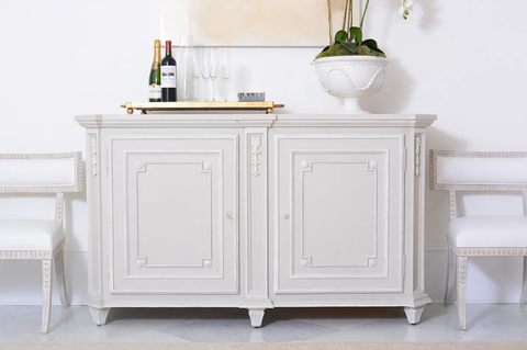 a side cabinet painted in a warm white, styled with a wooden board, bottles, and a fruit bowl, against a cool white wall