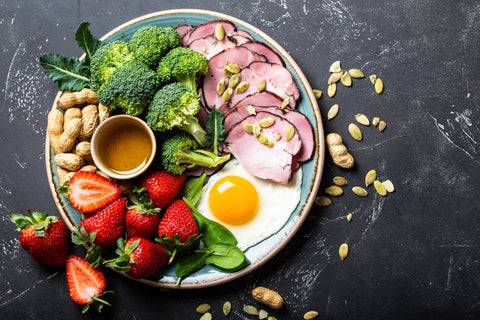 plate full of eggs, meat, strawberries, nuts, and broccoli on black background