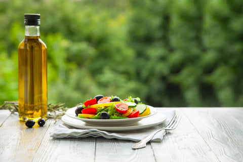 bottle of olive oil sits next to bright salad on white plate against green tree background