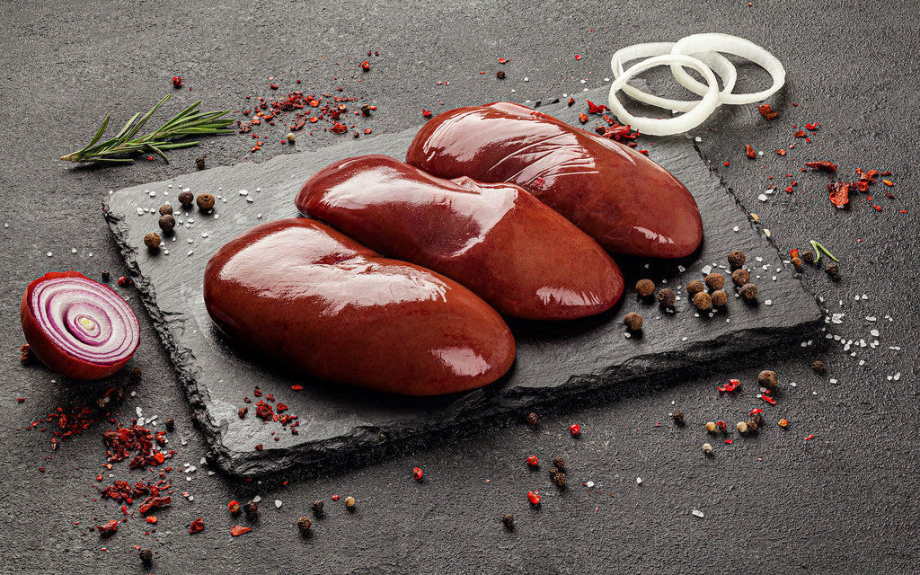 Three pieces of raw lamb kidney on a stone slab and surrounded by herbs and spices