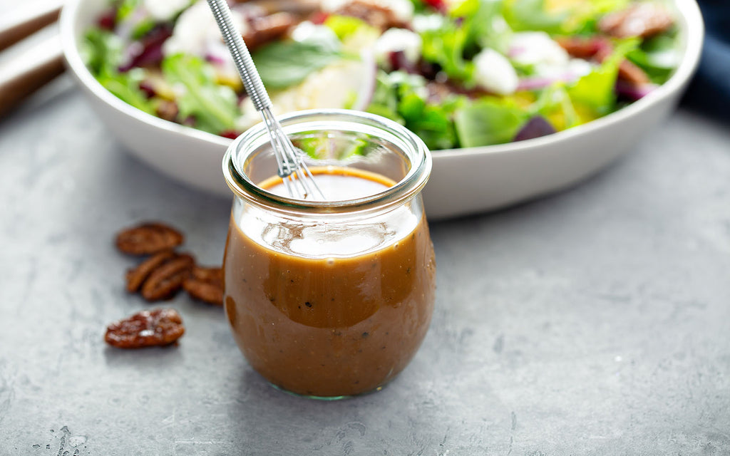 Salad dressing in a small glass jar with a whisk and a bowl of salad