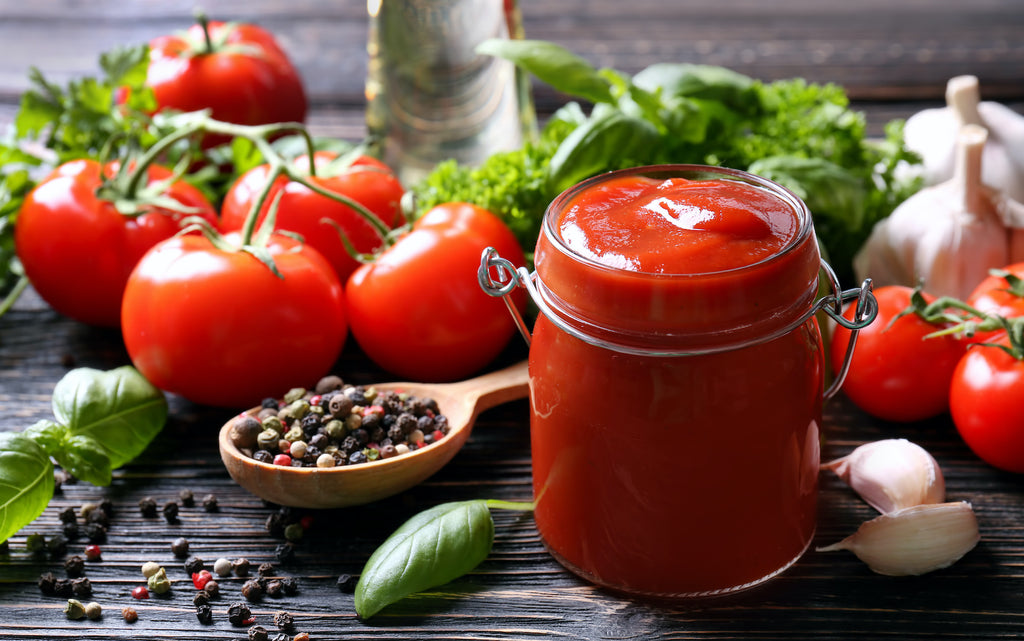 keto ketchup: Jar full of ketchup surrounded by tomatoes, herbs and spices on a wooden surface