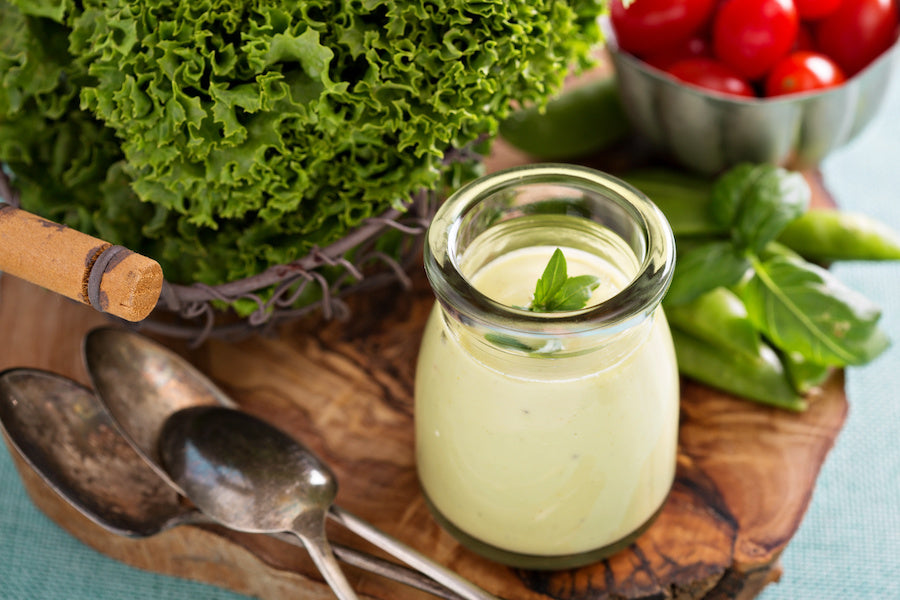 Mayonnaise in a small glass jar with green vegetables, tomatoes and spoons on top of a wooden board