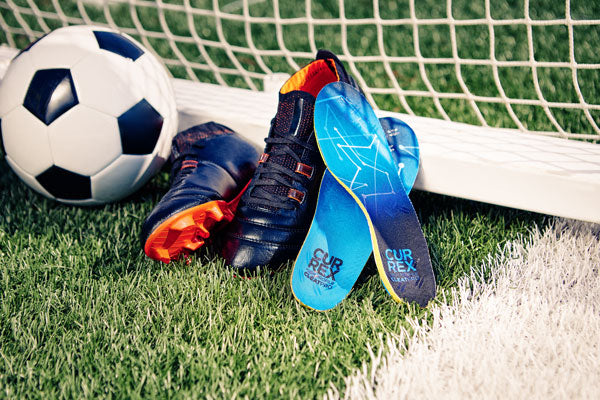 CURREX blue soccer insoles, soccer cleats, and soccer ball next to a goal net