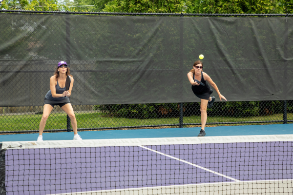 Two women playing pickleball while one serves behind the baseline