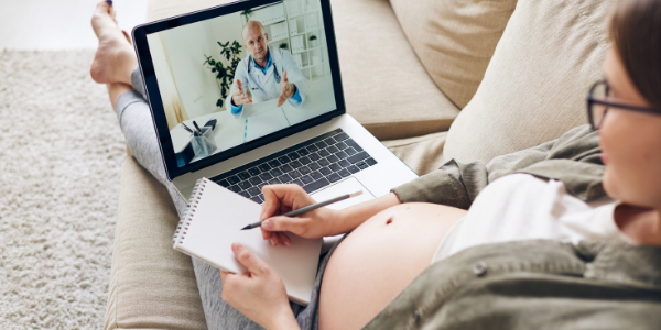 pregnant woman lying on the sofa and making notes while discussing with the doctor on tablet