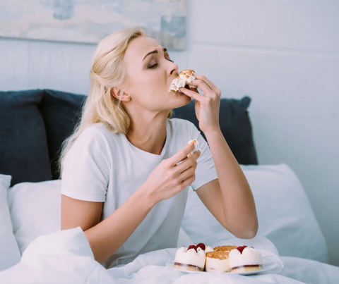 A woman in a white t-shirt sits on a bed, indulging in a large sandwich with visible enjoyment. Beside her is a plate with two more desserts, suggesting a moment of stress eating.