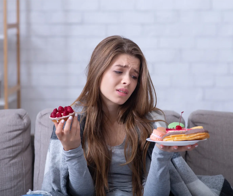 distressed young woman in a gray sweater sits on a couch, holding a piece of pie in one hand and a plate with a doughnut and éclair in the other, expressing regret or worry potentially related to stress eating