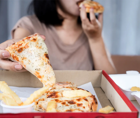 A woman in a casual brown top eats a slice of pizza with fries on the side, with another half-eaten pizza slice in the background, illustrating a possible case of eating in response to stress.