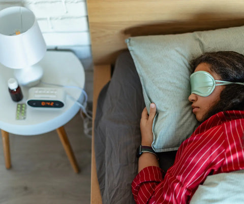 A person sleeping soundly with an eye mask on, a bottle of melatonin, and sleep-related items on a nightstand nearby.