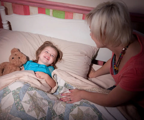 A young child in bed, laughing with delight as an adult female tucks them in for the night with a teddy bear by their side.