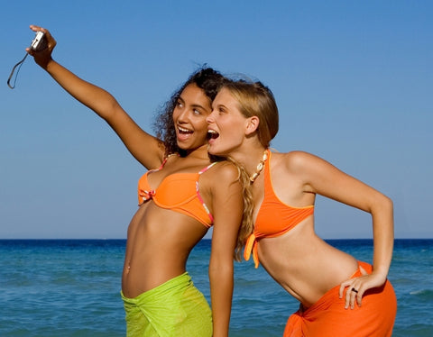 : Two tanned teenage girls in swimsuits taking a selfie at the beach.