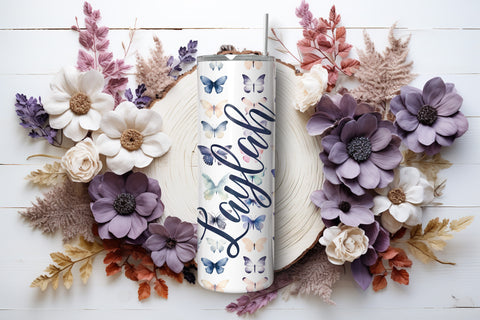 Butterfly skinny tumbler with a straw, laying on its side with flowers on either side.