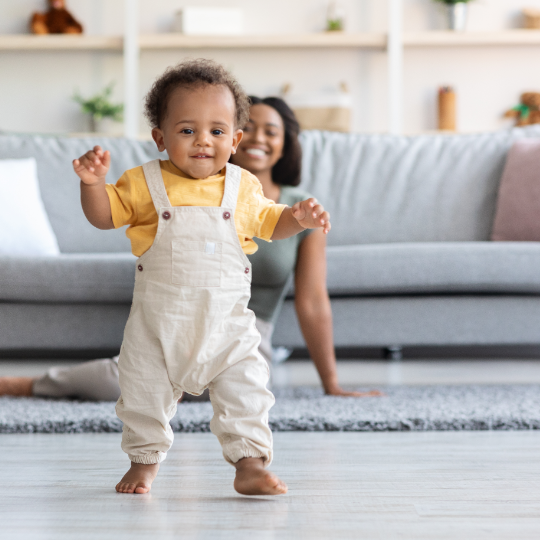 Happy baby confidently taking first steps in a babyproofed living room with caregiver watching from behind.