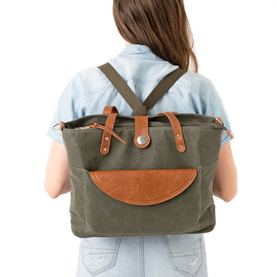 A woman in blue blouse and jeans shown from behind in front of a white background wearing a forest green waxed canvas backpack with brown vegan leather accents.