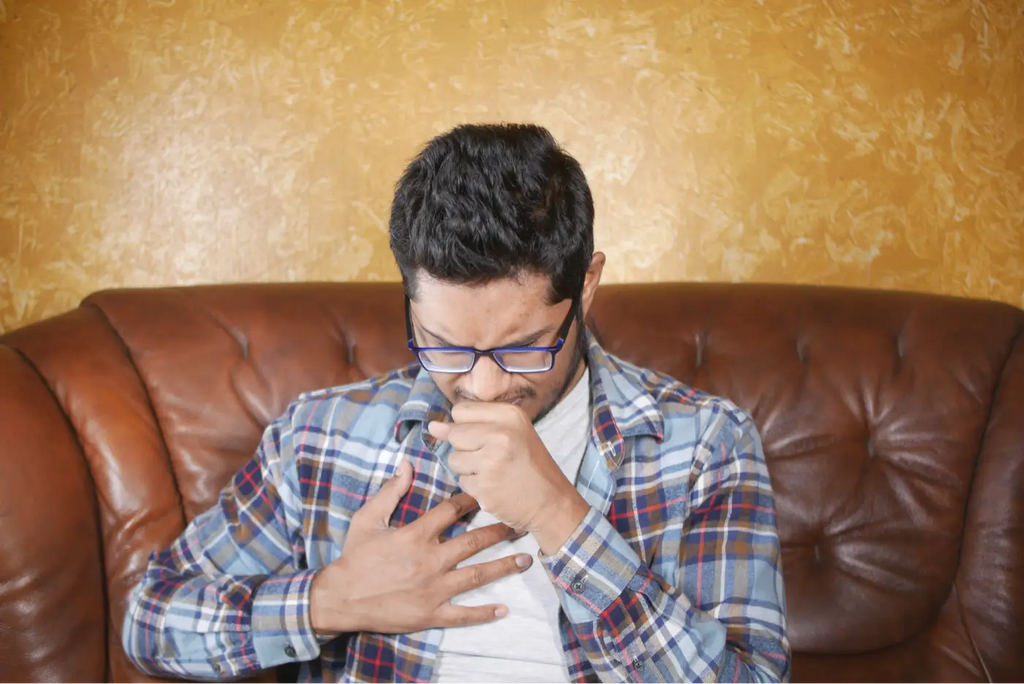 dark-haired man with glasses coughing sitting on leather couch