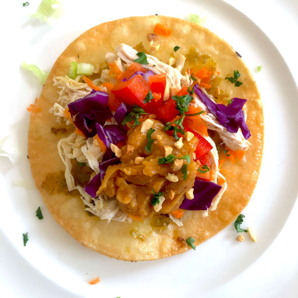 Birds eye view photo of colourful Mexican Chalupa with pulled pork, green salsa, purple cabbage, red bell pepper, peanut ginger sauce, peanuts, green onion, cilantro and carrots on top, on a white plate.