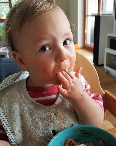 baby isst Spaghetti und ist im ganzen Gesicht und auf den Händen voller Sauce. auch die Biokleidung hat Flecken