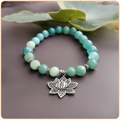 Dessin d'un bracelet de perles d'amazonite avec un pendentif plat et argenté en forme de fleur de lotus sur un sol gris et des feuilles vertes en fond Kaosix