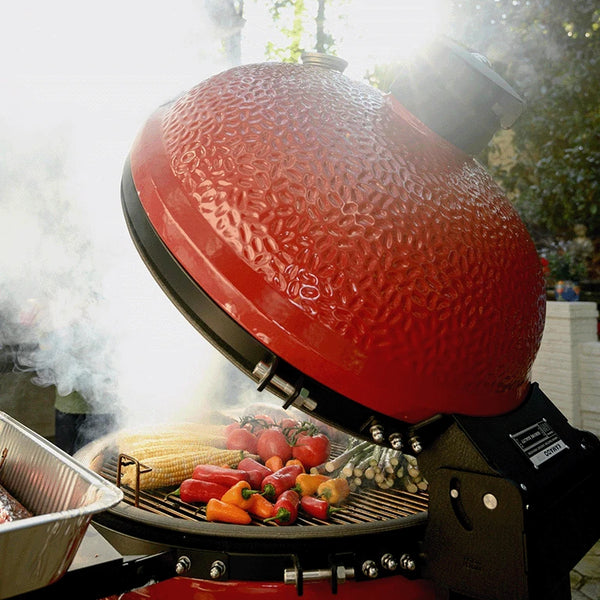 Smoke billows out of an open Kamado Joe grill filled with a variety of vegetables. The view shows the grill from the side and rear to show the Air Lift hinge.