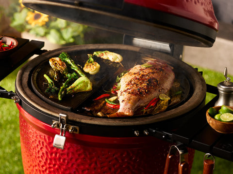 A whole fish grills on one side of a Kamado Joe grill while vegetables roast in a pan on the other side.