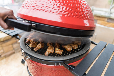 A person raised the lid of a Kamado Joe grill, showing a rack full of chicken wings cooking inside