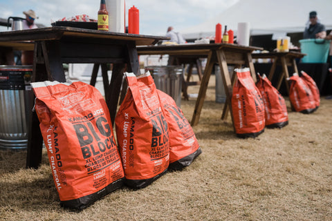 A row of tables set for serving barbecue outside. Bags of Kamado Joe Big Block lump charcoal are propped up against the table legs.