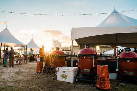 The sun on the horizon peeks through tents at a grilling competition. The nearest tent has 4 Kamado Joe grills with open bags of Big Block lump charcoal scattered between them.