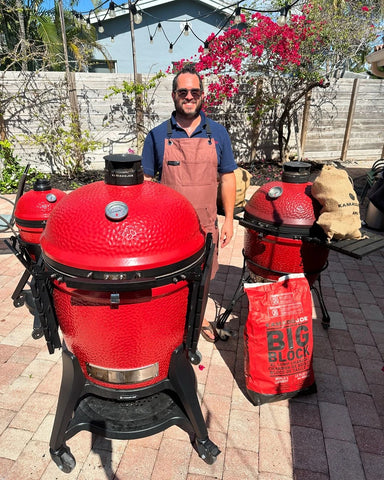 Oded Eschel wearing a blue, short-sleeved shirt and an apron stands between 3 Kamado Joe grills. Bags of Big Block charcoal and smoking wood are scattered around Oded and the grills.