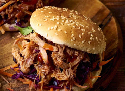 Closeup of a pulled pork sandwich on a sesame seed bun. The pork is topped with sauce and fresh cilantro, and rests on a mound of grated vegetables.