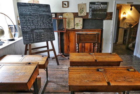 the image shows An old fashioned school room with some wooden desks and a blackboard that has some scribbles all over it