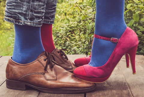 Some brown men's shoes with a men wearing some blue and pink socks and some pink, women's stilettos with the woman wearing blue socks