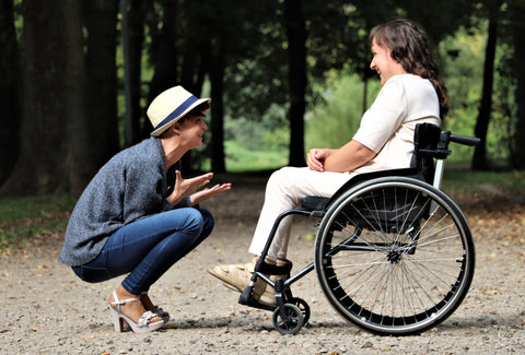 A lady in a wheelchair, in front of a group of trees, with another lady crouched in front of her – the pair are chatting