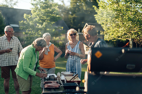 A group of men and woman are gathered around a bar-be-que in a sunlit garden; they are all talking happily