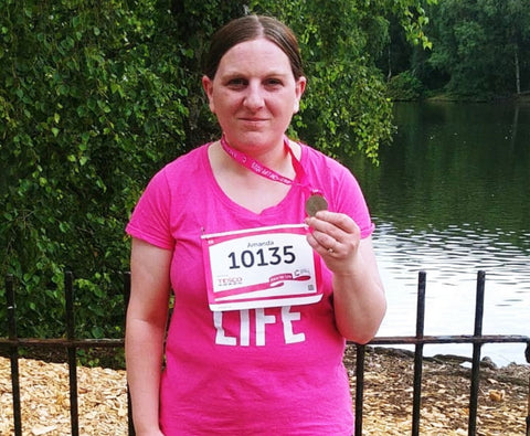Amanda Nicholson holding a Race for Life medal; she is wearing a pink running jersey and is standing in front of a pond 
