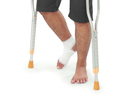 The lower half of a man; his right foot is bandaged and his left foot is without shoes and socks. There are two crutches that the man is using that are on either side of him