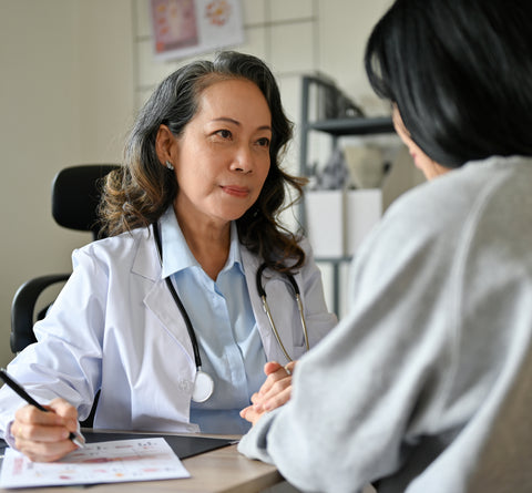 Two women sitting either side of a desk. One woman has a white, "medical" coat on and is wearing a stethoscope around her neck. The scene looks like a doctor talking to a patient 