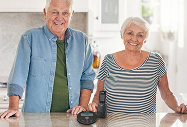 An elderly couple standing behind a work counter - they are both smiling and look very happy. The Call Blocker is on the work top