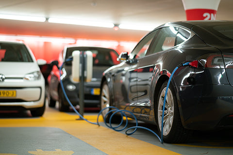 A number of electric cars in a garage. One black car is being charged by a cable from the car to an electricity charging point