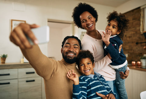 A man, woman and two children (male and female) are all standing in the kitchen. The man is holding out his mobile phone and the whole family are looking at it, smiling, as though they are talking to someone on the phone