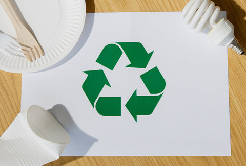 The recycling logo on a wooden table with a paper plate, plastic cup and a lightbulb