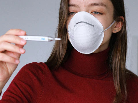 the image shows a woman wearing a mask checking her temperature on a thermometer