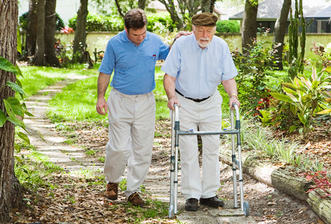 A man using a Zimmer frame on a country path, he is being helped by a friend