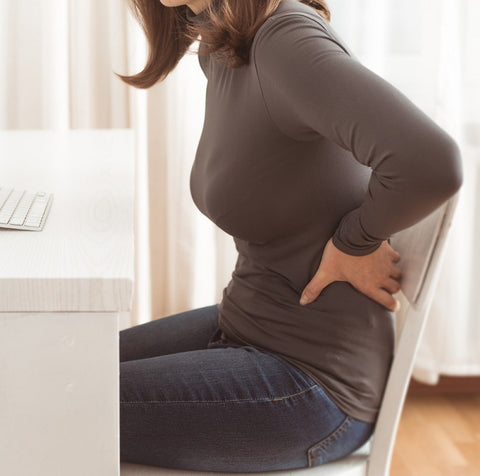 An image of a woman sat at a computer desk. She is holding her lower back, presumably from back pain due to posture.