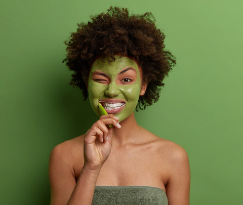 A woman with curly hair practicing self-care. She has a face mask on, a towel wrapped around herself and is brushing her teeth.