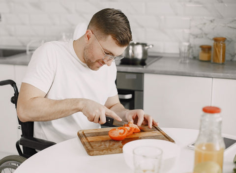A man in a wheelchair preparing vegetables for cooking. He is chopping up tomatoes on a wooden cutting board.