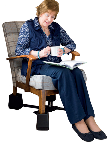  A woman sitting in a chair drinking a cup of tea and reading a book. Each leg of the chair has a chair raiser on it. The raisers have a cross bar connecting them all which is more secure than having four separate raisers