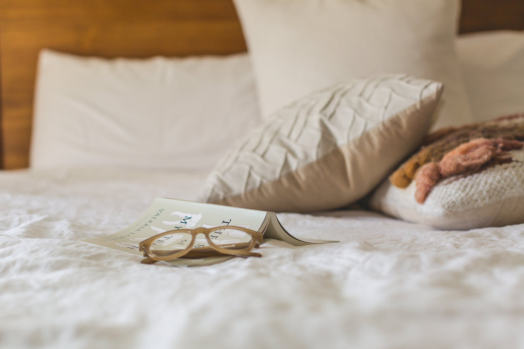 A bed with a blanket, pillow and a book and some glasses on it